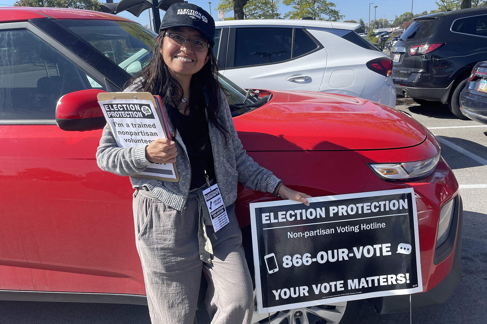 Volunteer with long dark brown hair wearing an election protection hat standing in front of a red sedan holding an Election Protection yard sign and clipboard. The volunteer is wearing an Election Protection lanyard that reads 